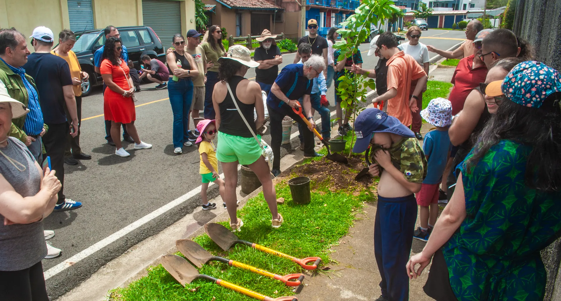 Vecinos de todas las edades reunidos en la calle durante la siembra del Día del Árbol, con palas listas sobre el zacate.